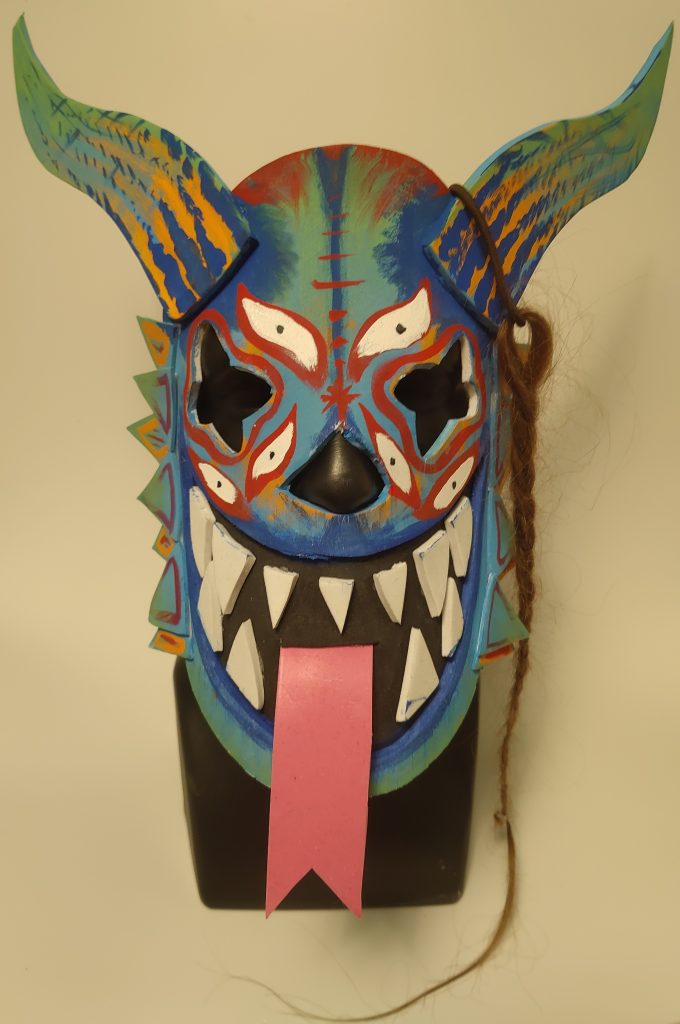 A mask grinning with sharp teeth and sticking out a forked tongue. It has two long horns curving up, spikes along the cheeks, and six additional eyes painted on the front. A braided lock of hair hangs from the left horn.